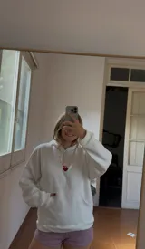 Canguro color crudo de algodón con felpa, de corte holgado y diseño con capucha. Presenta un bordado pequeño en el centro del pecho acompañado de un aplique de piedras en forma de corazón.