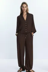 Pantalón negro de tiro alto con cintura elástica ajustable mediante cordones con terminales dorados. Presenta pernera ancha y bolsillos laterales. La tela tiene una textura sutilmente arrugada o plisada.