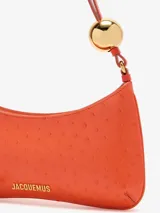 Cartera tipo bandolera Jacquemus Le Bisou Perle de cuero color naranja con efecto avestruz, de silueta curva y correa regulable con esfera metálica dorada. Incorpora el logotipo metálico de la marca en el frente y cierre superior.