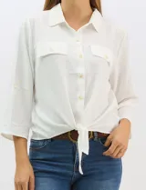 Camisa blanca de rayón con textura sutil, cuello con solapa, cierre frontal con botones y bolsillos simulados en el pecho. Presenta mangas 3/4 regulables con presilla y un diseño con nudo en el ruedo frontal.