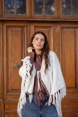 Poncho tejido de color blanco, con flecos en los bordes y detalles de bordado en crochet a lo largo de la abertura frontal. Cuenta con un cierre de cordones con borlas en el cuello.