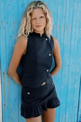 Top estilo chaleco de color azul marino, con cuello mao, sin mangas y trabillas en los hombros. Cuenta con cierre frontal cruzado mediante botones metálicos y bolsillos delanteros de plastrón con solapa.