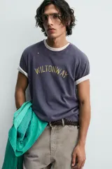 Camiseta de manga corta, corte holgado, color gris oscuro o azul marino con efecto lavado y rotos. Estampado frontal con el texto "WILTONWAY" en letras doradas.