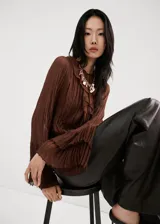 Blusa de color marrón chocolate, confeccionada en tejido plisado con textura. Presenta un diseño de cuello redondo con detalle de lazo, mangas largas amplias con puños acampanados y un corte holgado con volados sutiles en la parte frontal.