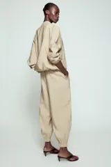 Pantalón satinado de tiro alto color beige, con cintura elástica ajustable con cordones, bolsillos delanteros y puños elásticos en los tobillos.
