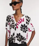 Blusa de viscosa con escote en V y mangas cortas abullonadas. Presenta un estampado de follaje y flores en tonos negro, fucsia y beige sobre fondo blanco.