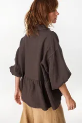 Camisa de lino marrón oscuro con silueta holgada, cuello camisero, cierre frontal de botones claros, mangas voluminosas y detalle de frunces en la cintura.