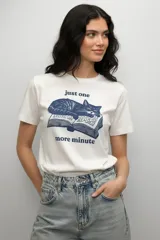 Remera de manga corta color blanco, con cuello redondo a la base y corte holgado. Presenta un estampado frontal en color azul que muestra un gato durmiendo sobre un libro abierto, acompañado por el texto 'just one more minute'.