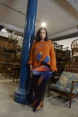 Sweater tejido a mano en lana merino color naranja, con bordado de texto en azul y apliques de uvas en azul. De calce holgado, cuello redondo y puños ajustados.