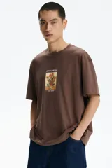 Remera marrón de algodón con cuello redondo, mangas cortas y hombros caídos, de corte holgado. Presenta un estampado frontal con la obra 'Los girasoles' de Van Gogh y el texto 'Sunflowers' en la parte superior y '-Van Gogh-' en la inferior.