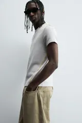 Camiseta de corte relajado, color gris claro, confeccionada en tejido con estructura waffle. Presenta cuello redondo y manga corta, con costuras decorativas en contraste color dorado que recorren los laterales y los hombros.