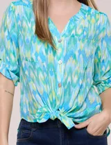 Blusa camisera de estilo casual confeccionada en mezcla de rayón y poliéster, con estampado multicolor en tonos turquesa, verde y celeste. Presenta escote en V con cuello mao, abotonadura frontal con botones nacarados, bolsillos aplicados en el pecho y mangas tres cuartos con presilla para arremangar. Cuenta con ruedo curvo y picos frontales diseñados para anudar en la cintura.