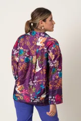 Blusa tipo casaca italiana abotonada, de satén estampado con fondo violeta y flores en colores azul, mostaza y naranja. Tiene cuello de camisa y mangas largas con presilla para sujetar.