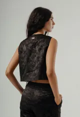 Top negro sin mangas, estilo chaleco corto, con cuello redondo y cierre frontal de botones. Presenta un tejido con textura jacquard de diseño floral o vegetal en tono oscuro.