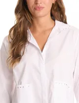 Camisa blanca de corte recto y estilo clásico, con cuello solapa, cierre frontal oculto y mangas largas con puños. Presenta dos bolsillos frontales decorados con apliques de tachas metálicas en el borde superior.