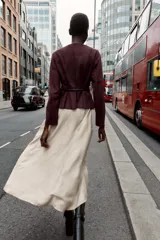 Chaqueta color beige con efecto ante, cuello redondo, manga larga y cierre frontal con cremallera. Presenta un bajo en evasé y un cinturón fino de cuero marrón oscuro con hebilla metálica.