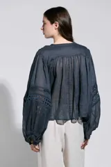 Camisa azul semitransparente con mangas abullonadas estilo globo, cuello redondo y cierre frontal con botones. Presenta detalles de encaje calado en las mangas.