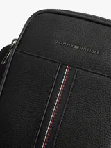 Morral de cuero sintético con acabado granulado color negro, de Tommy Hilfiger. Cuenta con un compartimento principal con cierre de cremallera, bolsillo frontal decorado con una franja tricolor vertical, correa cruzada ajustable y el logo metálico de la marca en la parte superior.