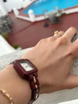 Reloj de pulsera con caja cuadrada y correa de eslabones de madera color borgoña. El dial es plateado con agujas y detalles dorados.