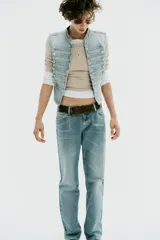 Chaleco de denim color celeste con cuello subido, sin mangas, con detalle de aplicaciones horizontales y botones metálicos en el frente. Cuenta con cierre frontal con ganchos metálicos ocultos.