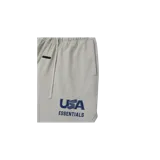 Short de felpa color gris claro, de corte holgado, con cintura elástica y cordón de ajuste largo. Cuenta con un pequeño parche rectangular en la parte superior y la inscripción "USA ESSENTIALS" estampada en azul marino en la pierna izquierda.