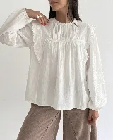 Blusa de algodón color blanco con cuello redondo, mangas largas con puños elásticos y detalles de puntilla y bordados en el frente y hombros.