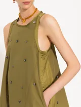 Vestido corto verde militar confeccionado en popelina, con cuello redondo y sin mangas. Presenta detalles de piedras y tachas metálicas en el delantero y la espalda. Las sisas, el escote y las aberturas laterales están terminadas con satén al tono. El acceso es mediante cierre metálico en la espalda.