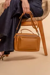 Bandolera de diseño rectangular en color suela, confeccionada en material sintético. Cuenta con un asa superior fija, correa larga ajustable y extraíble, y dos bolsillos frontales con cierre metálico, uno de ellos con diseño superpuesto. Presenta el logo de la marca grabado en el frente.