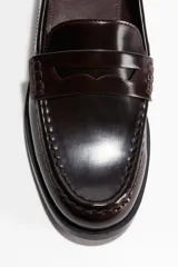 Mocasines tipo penny de cuero marrón oscuro (color borgoña/caoba) con acabado brillante y costura tradicional en el empeine. Suela y tacón bajos negros.