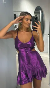 Vestido corto de tela metalizada en color violeta, con breteles finos, escote en V y aberturas laterales en la cintura. Presenta un diseño con volados en el ruedo.