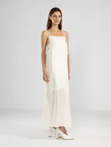 Vestido largo hasta el tobillo color blanco roto, con escote recto y finos breteles. Presenta un tejido liviano con textura arrugada y lazos ajustables en los costados.