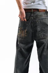 Pantalón de denim azul desgastado, corte mega baggy con piernas anchas y bolsillos traseros grandes. Presenta un bordado dorado en estilo gótico en el bolsillo trasero izquierdo.