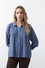 Camisa de manga larga color azul, con cuello camisero y cierre frontal de botones. Presenta un diseño con tablas verticales en la parte superior delantera y mangas con puños abotonados.