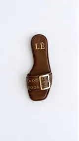 Sandalias planas color marrón con tira ancha decorada con ojales metálicos dorados y una hebilla grande, de diseño liviano y puntera cuadrada.