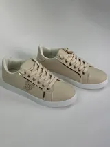 Zapatillas deportivas de cuero sintético en color blanco, beige y negro, con cordones y aplique decorativo dorado con forma de moño.