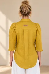 Camisa de lino color blanca, con cuello clásico, mangas tres cuartos abullonadas en los hombros y terminación con volados en los puños, cierre frontal con botones ocultos.