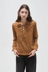 Blusa de gamuza sintética color camel, con cuello camisero y cierre frontal mediante cordones entrelazados. Presenta mangas largas y un corte holgado.