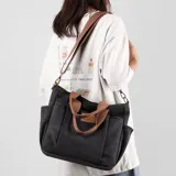 Bolso tipo tote bag de lona color negro, con múltiples bolsillos exteriores y asas cortas y correa larga de tela color marrón. Presenta un parche de cuero marrón con logo grabado en el frente.