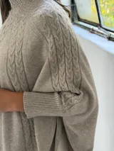 Poncho de tejido de punto color visón, con cuello alto y diseño de trenzas verticales. Presenta mangas cortas ajustadas y un corte holgado y redondeado en el ruedo.