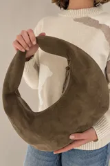 Cartera tipo morral confeccionada en gamuza color marrón chocolate, con diseño hobo de líneas curvas, asa integrada al hombro y detalles de tiras de cuero con placa metálica grabada.