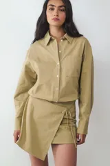 Vestido corto de algodón color beige, con diseño de camisa de manga larga en la parte superior y falda cruzada tipo pareo con bajo asimétrico y lazada lateral.