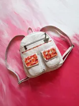 Mochila de cuero color blanco con diseño artístico pintado a mano en tonos naranjas y rojos en los bolsillos frontales. Cuenta con cierres metálicos YKK, herrajes de oro viejo y tiras ajustables.