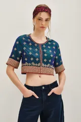 Camisa de seda con diseño tipo chaqueta corta, de color azul petróleo con bordados decorativos en tonos dorados y rojos. Presenta mangas largas abullonadas de color crema y cierre frontal.