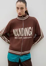 Campera tejida de punto color marrón con cierre frontal completo, cuello alto y detalles de rayas blancas en cuello, puños y ruedo. Presenta el logo de la marca estampado en el frente.