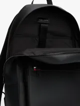 Mochila urbana Tommy Hilfiger de diseño minimalista en color negro. Presenta un compartimento principal amplio con cierre, un bolsillo frontal con cierre y detalle de franja tricolor distintiva de la marca. Incluye asa superior, correas ajustables acolchadas y logo metálico grabado en la parte superior.