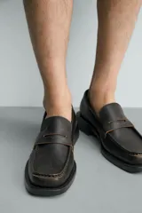 Mocasín de vestir color marrón oscuro, con empeine liso y detalle de antifaz. Presenta horma cuadrada y suela con ligero tacón geométrico a contraste.