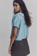 Blusa de viscosa color celeste con cuello Peter Pan fruncido y ribete de encaje. Presenta cierre frontal con tres tiras para anudar, mangas cortas abullonadas y puños con elástico.