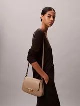Cartera mediana de poliuretano color tostado, con diseño estructurado, solapa frontal, cierre de broche a presión metálico dorado con el logo de Calvin Klein y correa ajustable.