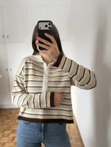 Sweater tipo chomba de tejido bremer, con diseño de rayas horizontales en tonos beige, blanco y marrón chocolate. Presenta cuello polo con cierre frontal de botones y mangas largas.