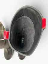 Bota de caña alta en cuero negro con punta redonda y taco bajo. Presenta un panel lateral de gamuza con bordado geométrico en hilo gris, herraje metálico decorativo en el empeine y cierre interno. El diseño cuenta con una boca asimétrica y forro interior de cuero.